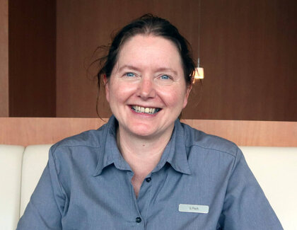 The dark-haired gastronomic manager Gabriele Pioch sits at a dark table with an anthracite runner and smiles happily into the camera. On the table are two cappuccinos and two brownies on a white plate and a green plant.