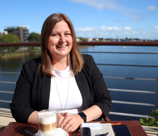 Die dunkelhaarige Ann-Christine Janzen sitzt glücklich auf der Hotelterrasse an einem braunen Holztisch. Auf dem Tisch vor ihr stehen zwei Latte Macchiato. Zudem liegt ein Notizbuch, ein mobiles Telefon, ein Handy, Visitenkarten und ein Schlüssel auf dem Tisch.