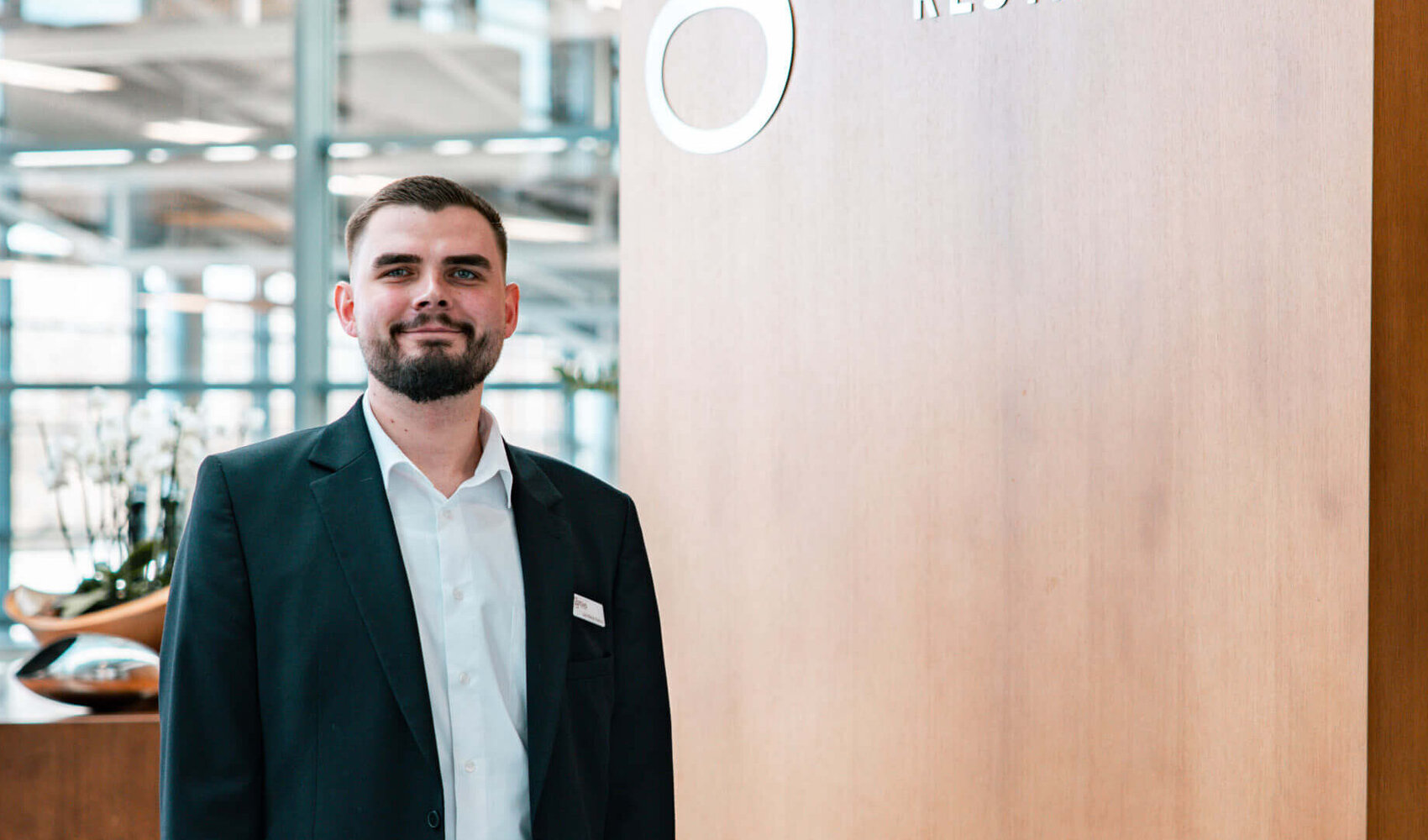 ATLANTIC success story – Deputy restaurant manager Jan Niklas Rakoci Dark-haired Jan Niklas Rakoci stands in front of a large wooden sign that reads ‘Gottlieb Restaurant’. He is wearing a white shirt, black trousers and a black jacket.