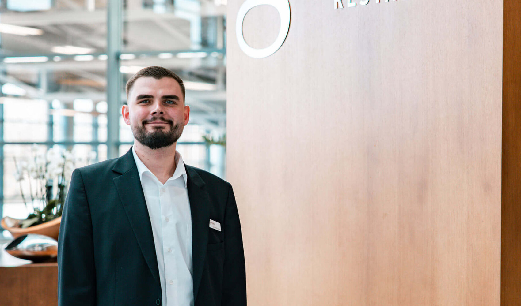 ATLANTIC Erfolgsgeschichte – Stellvertretender Restaurantleiter Jan Niklas Rakoci Der dunkelhaarige Jan Niklas Rakoci steht vor einem großen Schild aus Holz auf welchem "Gottlieb Restaurant" steht. Er trägt ein weißes Hemd, eine schwarze Hose und ein schwarzes Sakko.