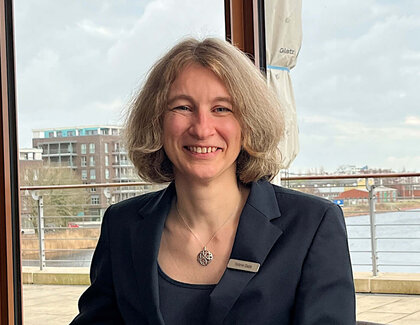 Empfangsleiterin Stefanie Gladis lächelt in die Kamera währen sie an einem dunklen, runden Restauranttisch sitzt. Im Hintergrund ist das Wilhelmshavener Hafenbecken zu sehen, vor ihr steht ein Cappuccino mit einem Brownie auf der Untertasse. Auch ihre Schlüssel liegt auf dem Tisch.