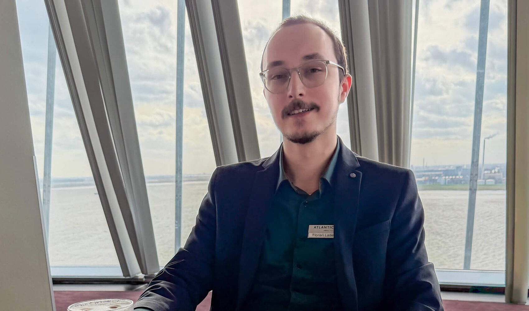 Der dunkelhaarige Florian Ladewig sitzt an einem hellen Holztisch sitzt und schaut in die Kamera. Eine Hand liegt auf dem Tisch, die andere auf seinem Bein. Ein Latte Macchiato steht und eine Schlüsselkarte sowie ein Chip-Schlüssel liegen vor ihm auf dem Tisch.  