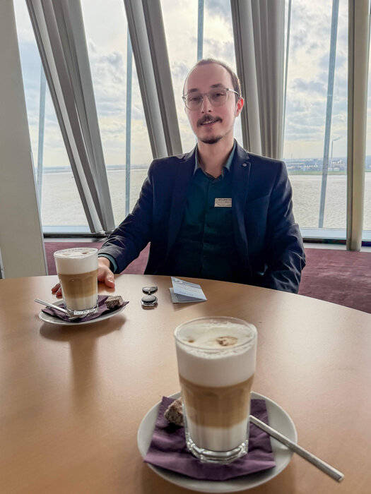 Der dunkelhaarige Florian Ladewig sitzt an einem hellen Holztisch sitzt und schaut in die Kamera. Eine Hand liegt auf dem Tisch, die andere auf seinem Bein. Ein Latte Macchiato steht und eine Schlüsselkarte sowie ein Chip-Schlüssel liegen vor ihm auf dem Tisch.  