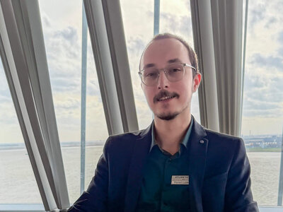 Florian Ladewig, with his dark hair, is sitting at a light wooden table and looking into the camera. One hand rests on the table, the other on his leg. A latte macchiato stands before him, and a key card and a key fob lie on the table in front of him.  