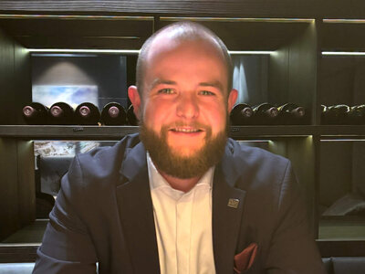 Der bärtige Leon Decker sitzt lächelnd im Weinkeller an einem Holztisch. Auf dem Tisch vor ihm steht ein Kaffee Crema. Seine Hände liegen gefaltet auf dem Tisch, daneben ein Durand-Korkenzieher.