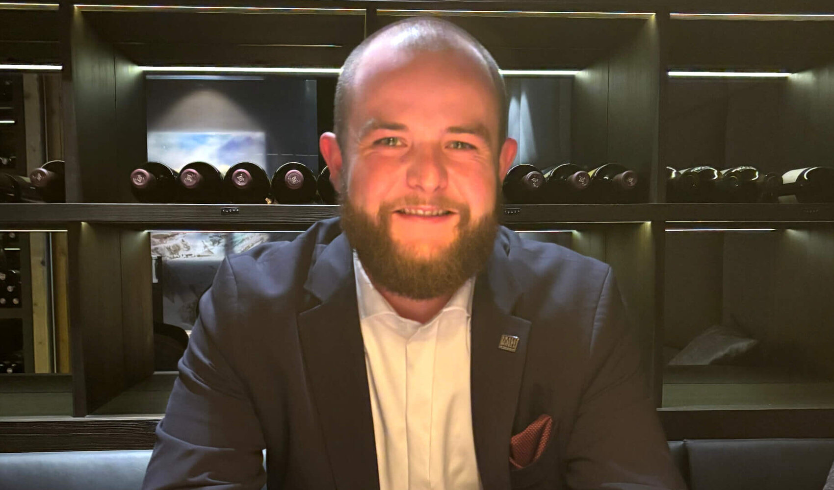 Auf einen Kaffee mit Restaurantleiter Leon Decker (Severin*s – The Alpine Retreat) Der bärtige Leon Decker sitzt lächelnd im Weinkeller an einem Holztisch. Auf dem Tisch vor ihm steht ein Kaffee Crema. Seine Hände liegen gefaltet auf dem Tisch, daneben ein Durand-Korkenzieher.