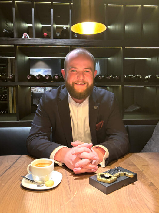 Der bärtige Leon Decker sitzt lächelnd im Weinkeller an einem Holztisch. Auf dem Tisch vor ihm steht ein Kaffee Crema. Seine Hände liegen gefaltet auf dem Tisch, daneben ein Durand-Korkenzieher.