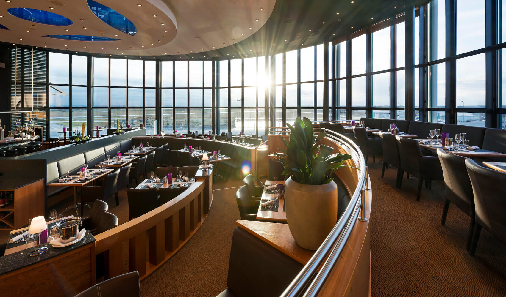 Das Restaurant BLIXX im ATLANTIC Hotel Airport in Bremen hat eine große Fensterfront, durch welche viel Sonne hereinkommt. Im Restaurant stehen viele Stühle und eingedeckte Tische sowie eine große Pflanze.